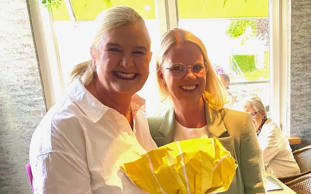 Frau Kaufhold lächelt mit einer Kollegin, beide halten ein gelbes Präsent – freundlicher Moment am Fenster.