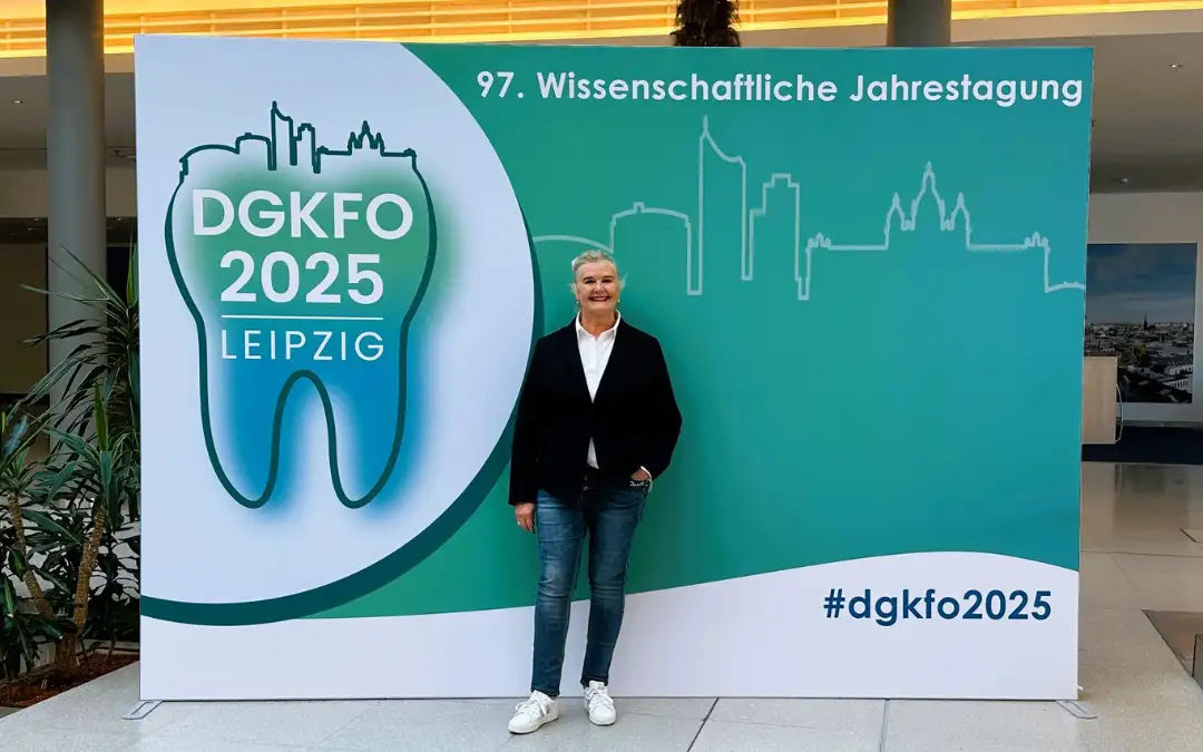 Iris Kaufhold steht vor dem großen Banner der 97. Wissenschaftlichen Jahrestagung der DGKFO 2025 in Leipzig. Das Logo zeigt einen stilisierten Zahn mit Skyline und Schriftzug „DGKFO 2025 Leipzig“.
